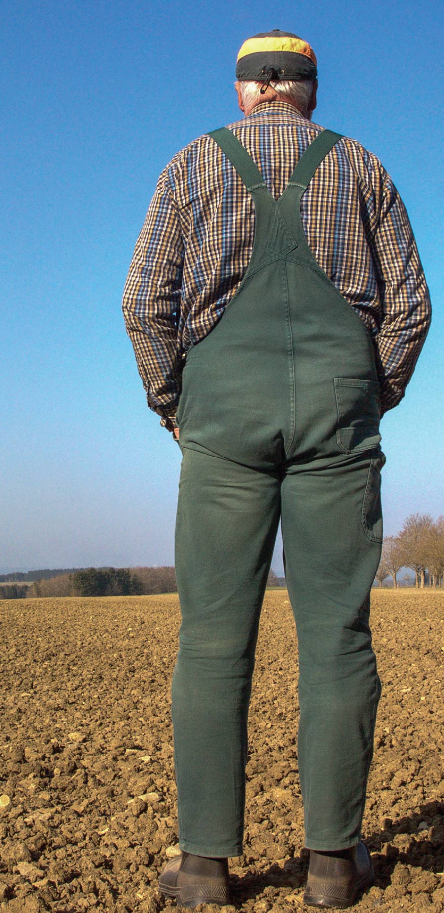 man in green overalls with plaid shirt standing in a field with back towards camera