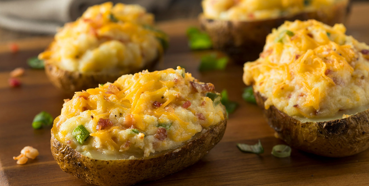 A landscape close-up photographic perspective of baked potatoes covered with cheese, bacon bits, and other assorted food toppings