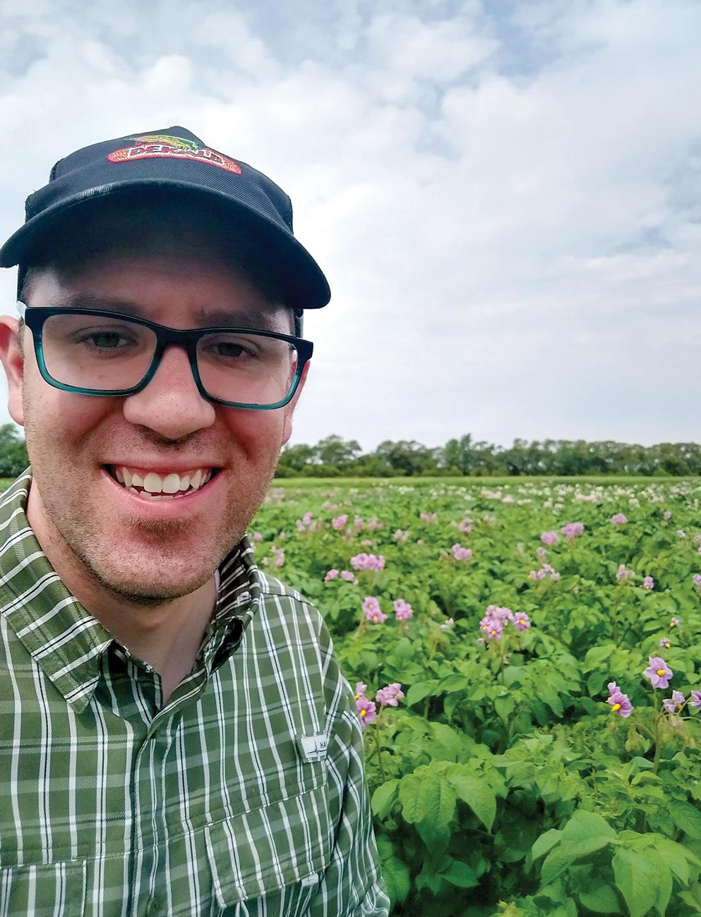 researcher Jed Grow takes a smiling selfie in a field of crops topped with purple flowers